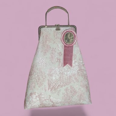 Rosa Victoriana Bolso de mano blanco con detalles rosados y un pin decorativo en una superficie rosa.